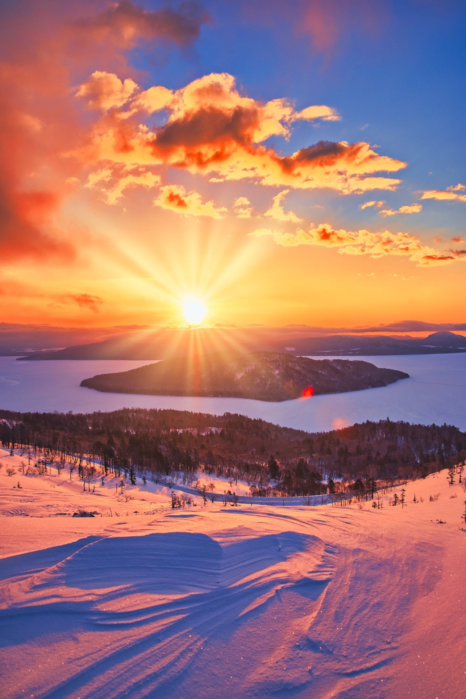 Atardecer invernal sobre un lago congelado en Hokkaido, con el sol brillando intensamente y cielo teñido de tonos naranja y rosa.